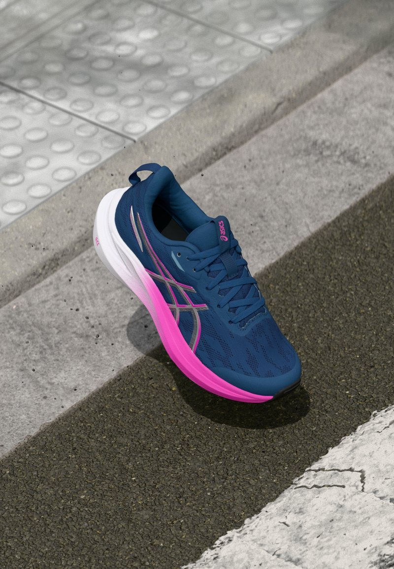 Chaussure de sport bleue avec une tige en mesh respirant, semelle rose et blanche, et accents réfléchissants, présentant un design rembourré pour le confort.