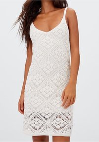 Robe blanche sans manches en dentelle complexe avec un motif géométrique. Présente un décolleté en V et une silhouette ajustée, se terminant au-dessus du genou.