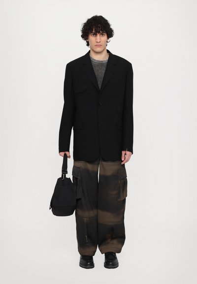 Black tailored blazer, gray textured top, oversized cargo pants with brown patterns, and black footwear. Model holds a small bag.
