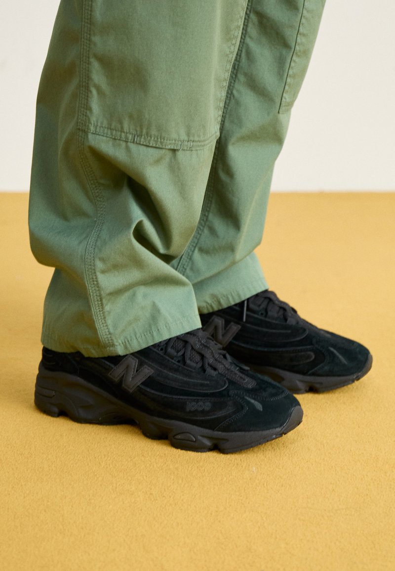 Zapatillas New Balance negras con un upper texturizado, que presenta un logotipo prominente y una suela acolchada, combinadas con pantalones verdes holgados.