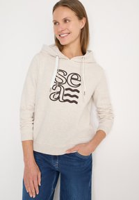 Hoodie beige clair avec une poche kangourou et des cordons blancs. Présente une impression noire du mot "sea" avec des accents ondulés.