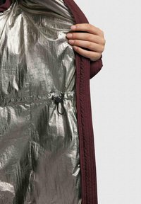 Burgunderfarbige Jacke mit Innenfutter aus reflektierendem Silbermaterial; ausgestattet mit einem Zugband mit Kordelstopper zur Anpassung der Passform. Strukturierte äußere Schicht.