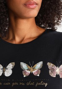 Camiseta de algodón negra con tres estampados de mariposas coloridas y detalles dorados, con el texto debajo que dice "me puedes dar esa sensación".