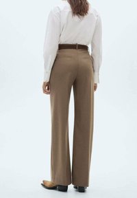 Pantalon marron sur mesure à coupe droite, avec ceinture et poches, associé à une chemise blanche à manches longues. Texture lisse.