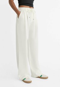 Witte wijde broek met plooien aan de voorkant, een tailleband met trekkoord en zijzakken. Gecombineerd met beige sneakers met groene accenten.