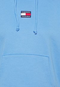 Blå huvtröja i mjuk bomull med en framficka. Har en patch-logotyp med "TOMMY JEANS" och ett röd, vit och blått randigt mönster.