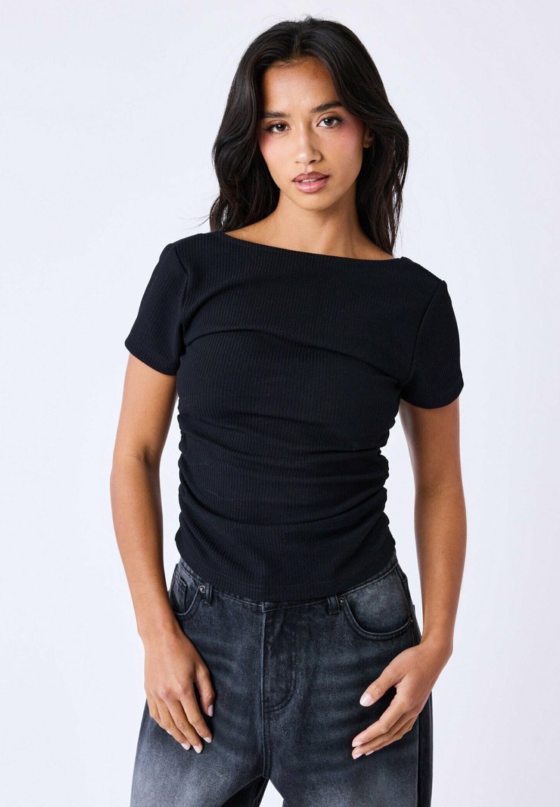 Haut noir à côtes avec des manches courtes, présentant un décolleté large et des détails froncés à l'avant. Associé à un jean noir délavé.
