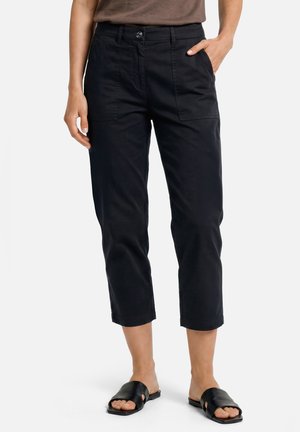 Vrouw draagt zwarte korte broek met voorop aangebrachte zakken en zwarte open-toe slidesandalen, staand met één hand in de zak.
