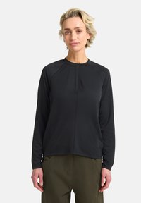 Schwarzes Langarmshirt mit Raglanärmeln, Rundhalsausschnitt und dezenten Faltendetails vorne, aus glattem Stoff gefertigt. Kombiniert mit grünen Hosen.