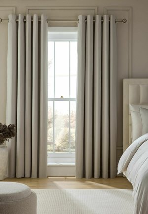 Floor-length beige curtains hang on a window in a neutral-toned bedroom with a bed, white rug, and a plant in a pot.
