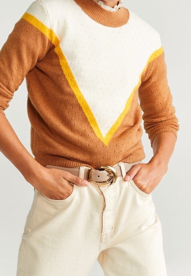 Personne les mains dans les poches portant un pull marron et blanc avec des accents jaunes et un pantalon beige clair avec une boucle de ceinture beige.