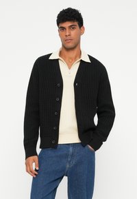 Zwarte gebreide cardigan met textuurpatroon, knoopsluitingen en geribbelde manchetten. Draag over een beige trui met ritssluiting aan de hals, in combinatie met blauwe jeans.