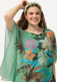 Grøn, gennemsigtig blomsterbluse med levende tropiske designs i orange, pink og blå. Løs pasform med brede ærmer og rund halsudskæring.