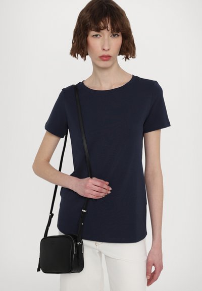 T-shirt bleu marine à manches courtes avec un col rond, associé à un sac bandoulière noir compact avec une longue sangle. Pantalons blancs.
