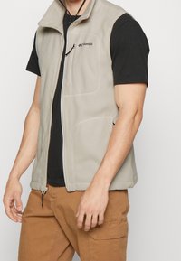 Ljusgrå fleeceväst med ståkrage, framdragkedja och synlig logotyp. Har två sidofickor och en justerbar nederkant för passform.