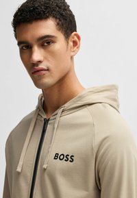Sudadera con cremallera en color beige, con cordones, que presenta el logo negro "BOSS" en el pecho, textura suave y un diseño de corte relajado.