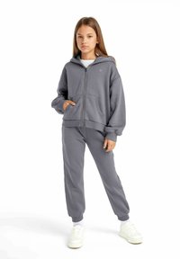 Grå zip-hoodie med stora framfickor, ribbade manschetter och matchande träningsbyxor. Vita sneakers kompletterar outfitens avslappnade stil.