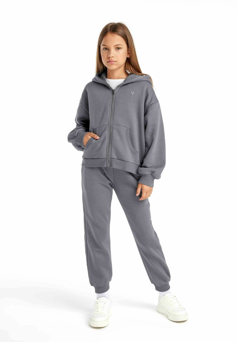 Grå zip-hoodie med stora framfickor, ribbade manschetter och matchande träningsbyxor. Vita sneakers kompletterar outfitens avslappnade stil.