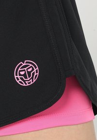 Černé sportovní šortky s růžovým logem, s obloukovým lemem a bočním rozparkem, nošené přes růžové kompresní šortky. Hladká textura látky.