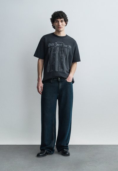 Νέος άνδρας με σγουρά μαλλιά φορώντας μαύρο γραφικό μπλουζάκι "Infinite Secret Tour '99", φαρδιά σκούρα τζιν και μαύρα παπούτσια, στέκεται μπροστά σε έναν γκρι τοίχο.