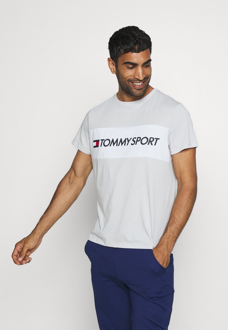 T-shirt grigio a maniche corte con un pannello bianco che presenta "TOMMYSPORT" in nero. Il tessuto sembra morbido; il modello lo ha abbinato a pantaloni blu.