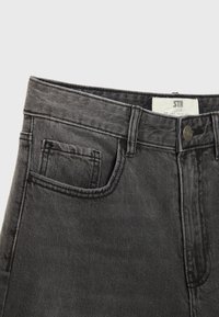 Jean en denim noir taille haute avec poche avant, passants de ceinture, fermeture à bouton et une étiquette blanche de marque visible à l’intérieur de la ceinture.