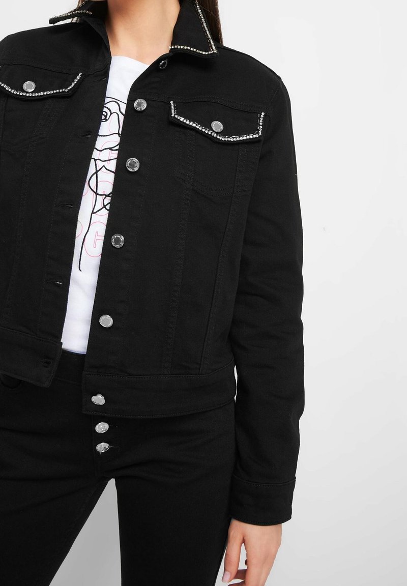 Black denim jacket with silver studded collar and pocket trims, featuring metal buttons and a tailored fit. Worn over a white graphic tee.