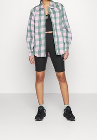 Person som bär en grön och rosa rutig skjorta över en svart Adidas crop top och cykelshorts, med svarta sneakers och ljusgröna strumpor.