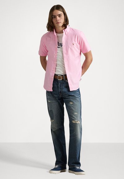 Chemise en coton rose clair à manches courtes, sur un t-shirt blanc avec du texte bleu marine, associée à un jean bleu déchiré et des chaussures en toile bleues.