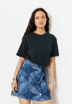 Femme aux longs cheveux foncés portant un T-shirt noir uni et une jupe en denim boutonnée avec de grands motifs géométriques.