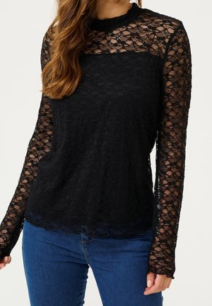 Femme portant un haut en dentelle noir à manches longues avec un motif floral, associé à un jean bleu, montré du cou jusqu’aux hanches.