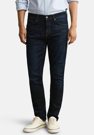 Mann trägt dunkelblaue Slim-Fit-Jeans, hellblaues hochgekrempeltes Hemd und cremeweiße Sneakers, steht vor einem schlichten Hintergrund.