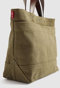 Beige plátěná taška s hnědými koženými uchy, přední kapsou a červenou štítkovou nálepkou Levi's v horním rohu.