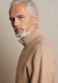 Beżowy sweter o teksturowanym splocie z wysokim kołnierzem, z subtelnym wzorem i luźnym krojem, przedstawiony z boku na neutralnym tle.