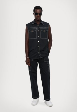 Zwarte mouwloze shirt met twee voorzakken en contrasterende witte stiksels, gecombineerd met bijpassende zwarte broeken en witte sneakers.