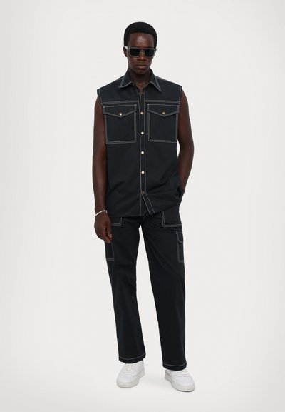Camiseta negra sin mangas con dos bolsillos frontales y costuras blancas en contraste, combinada con pantalones negros a juego y zapatillas blancas.