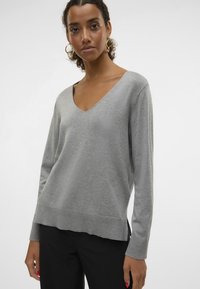 Pull gris en V en matériau doux, avec des manches longues et un ourlet légèrement arrondi. Associé à un pantalon noir.