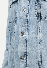 Ανοιχτό μπλε τζιν μπουφάν με εμφανείς μεταλλικά κουμπιά και λεπτομερή ραφές στο μπροστινό μέρος και τις τσέπες.