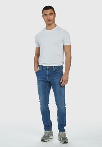 Vyriški mėlyni džinsai su slim fit kirpimu ir blukimo detalėmis, derinami su balta marškinėlių tipo maikė ir pilkais sportbačiais. Paprastas, kasdieniškas aprangos rinkinys.