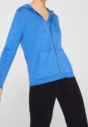 Osoba ubrana w niebieską bluzę z zamkiem błyskawicznym i przednimi kieszeniami oraz czarne spodnie sztruksowe na jednolitym tle.
