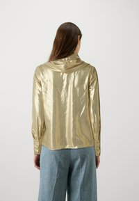 Camicia lunga dorata lucida con colletto drappeggiato, polsini con bottoni e taglio dritto, abbinata a pantaloni larghi blu chiaro.