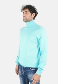 Maglione dolcevita turchese in materiale a maglia liscia con collo e polsini a coste. Indossato con jeans in denim blu.