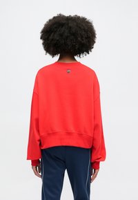 Červená mikina s dlhými, voľnými rukávmi a rebrovaným lemom. Okrúhly výstrih, na zadnej strane s malou nášivkou. Mäkko štruktúrovaná látka.