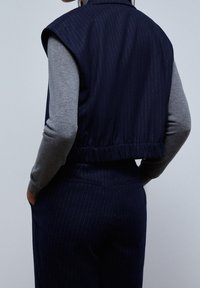 Navy berjalur cropped vest dengan leher bulat dan detail bahu, dikenakan di atas atasan lengan panjang abu-abu. Tekstur kain yang halus.