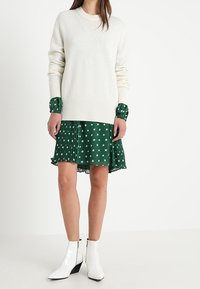 Biały sweter z dzianiny noszony na zielonej sukience w gwiazdy z plisami, uzupełniony błyszczącymi białymi botkami do kostki z geometrycznym obcasem.