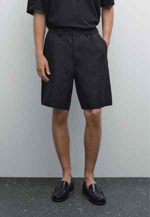 Black linen shorts with a relaxed fit, front pockets, and a button closure, paired with black loafers on a textured gray surface.