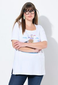 T-shirt en coton blanc avec un imprimé d'un oiseau portant une écharpe rayée sur un fond de ciel bleu clair, associé à un jean bleu foncé.