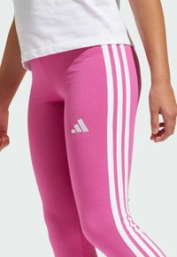 Leggings rosa realizzati in materiale elasticizzato con tre strisce bianche lungo i lati e un piccolo logo Adidas bianco sulla coscia sinistra.