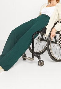 Green wide-leg trousers made of flowing fabric, paired with a white top and cream cardigan. The wearer is seated in a black wheelchair.