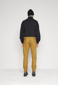 Svart tröja med hög krage, lös passform. Khakifärgade byxor med bakfickor. Svart mössa. Svarta stövlar. Neutral bakgrund.